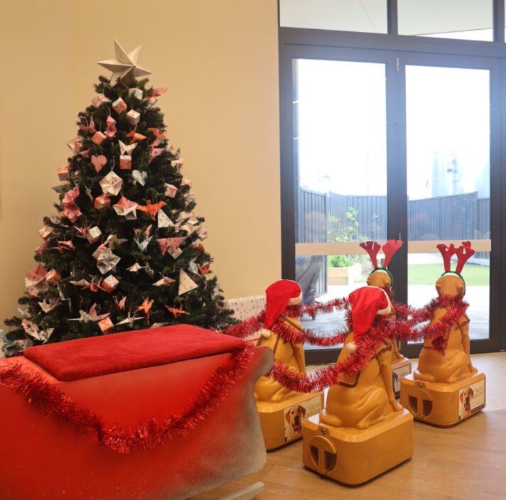A Santa sleigh led by four donation dogs, decorated with tinsel, Santa hats and reindeer ears. Behind the sleigh is a Christmas tree with origami ornaments.
