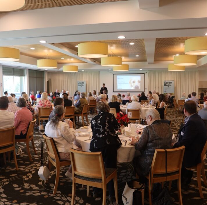 A large open room, set up in a gala style. Seated people at several round tables are pictured facing the front of the room.