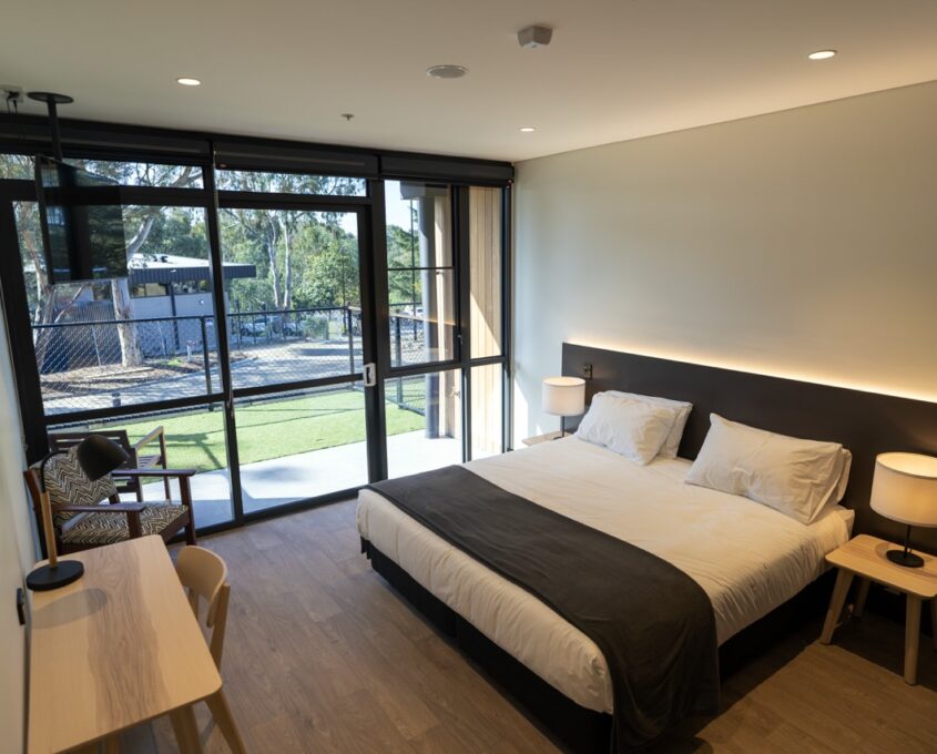 One of the residential rooms with a bed, floor to ceiling windows and a desk positioned opposite of the bed.