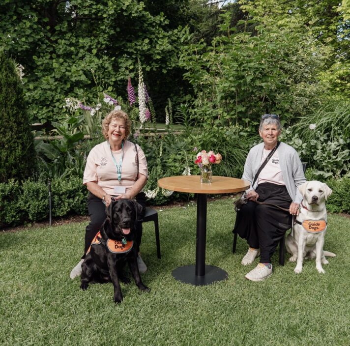 Two volunteers Puppy Raisers sitting at a table with the pups in their care. They are pictured in the beautiful gardens of Cranlana.