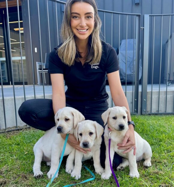 Puppy Development Advisor, Alex kneeling with three yellow Labrador puppies in the free run area. Alex is pictured smiling at the camera.