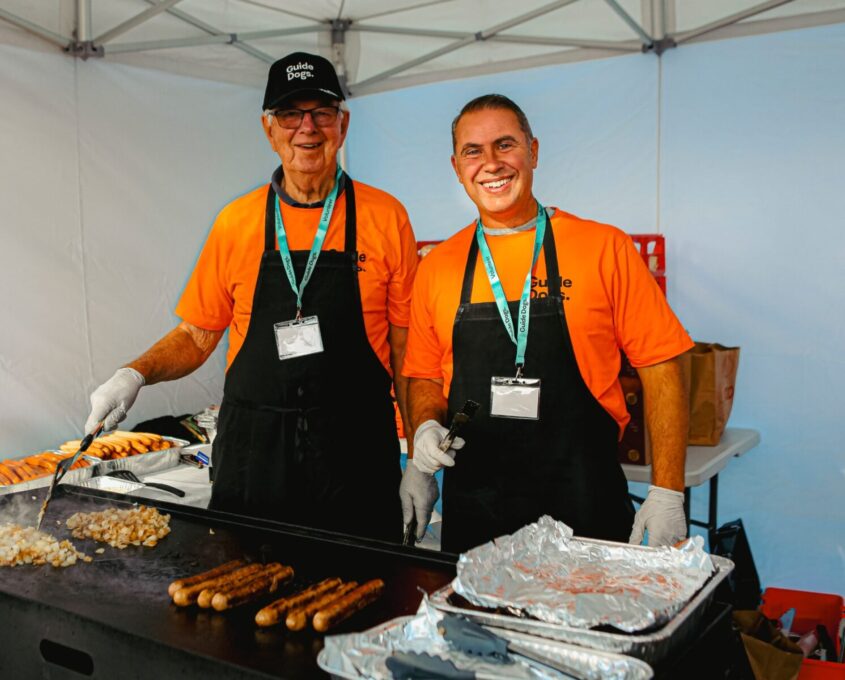 Two volunteers behind a BBQ cooking sausages.