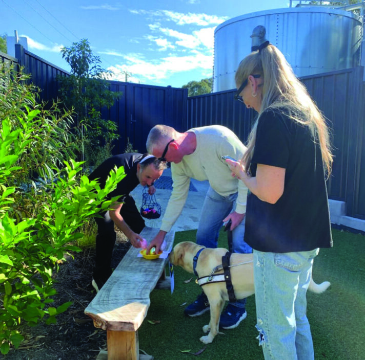 Three people and a guide dog gather around a bench outdoors, participating in an easter egg hunt.