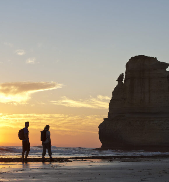 Two people with backpacks stand on a beach at sunset, facing a large rock formation in the water.