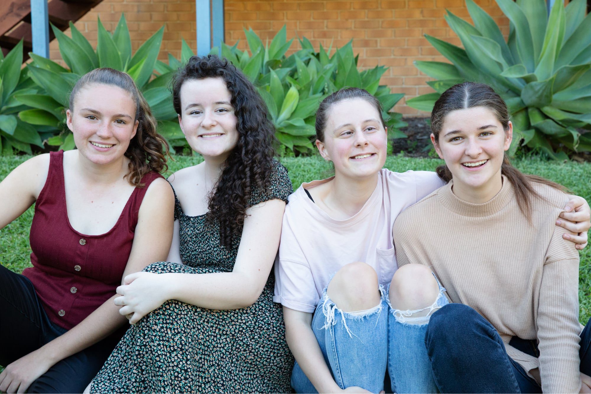 A group of four young adults sitting outside on the grass all in a row. Everyone is looking at the camera smiling.