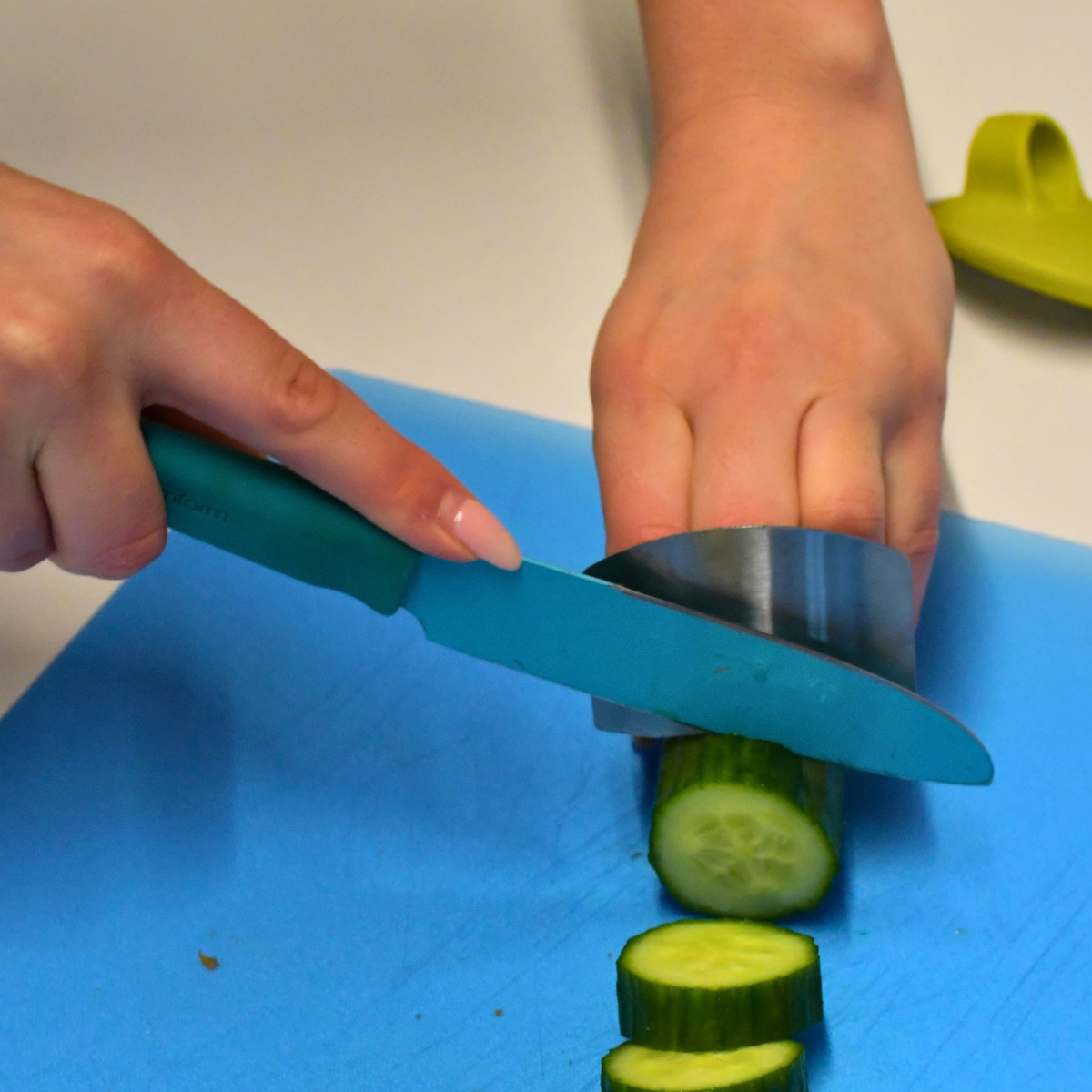 A person cutting a cucumber on a blue chopping board using a knife and knife guard blocking their fingers.