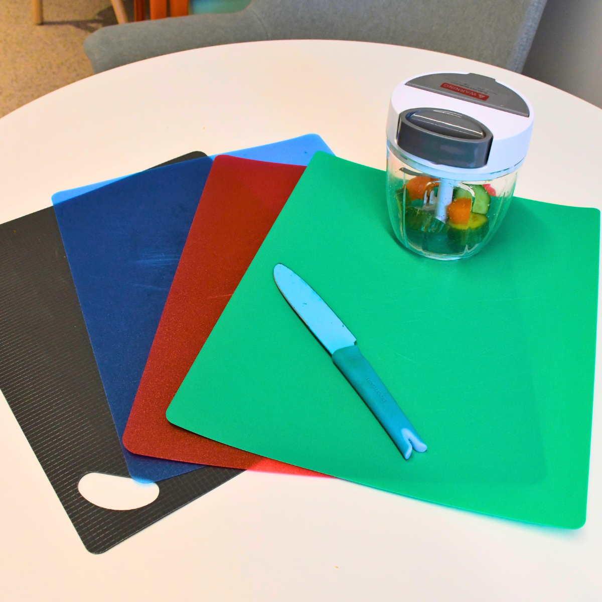 A selection of brightly coloured chopping boards, a kitchen knife and a manual food chopper arranged on a table.