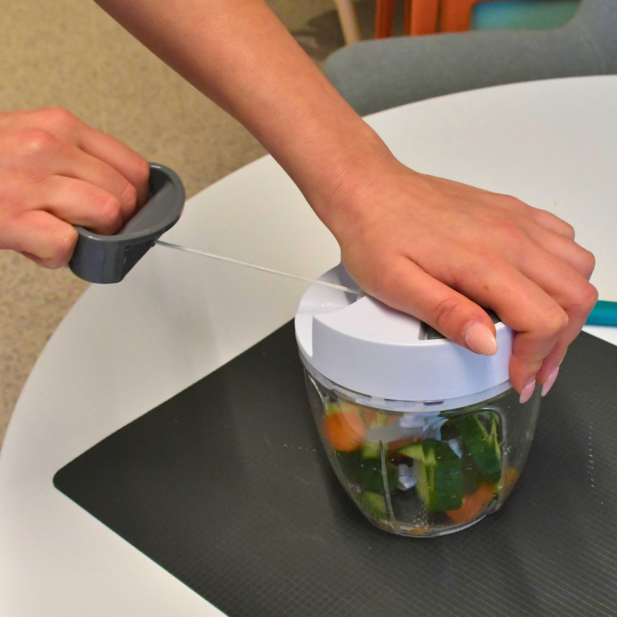A person using a manual food chopper containing chopped cucumber and carrot on a table.