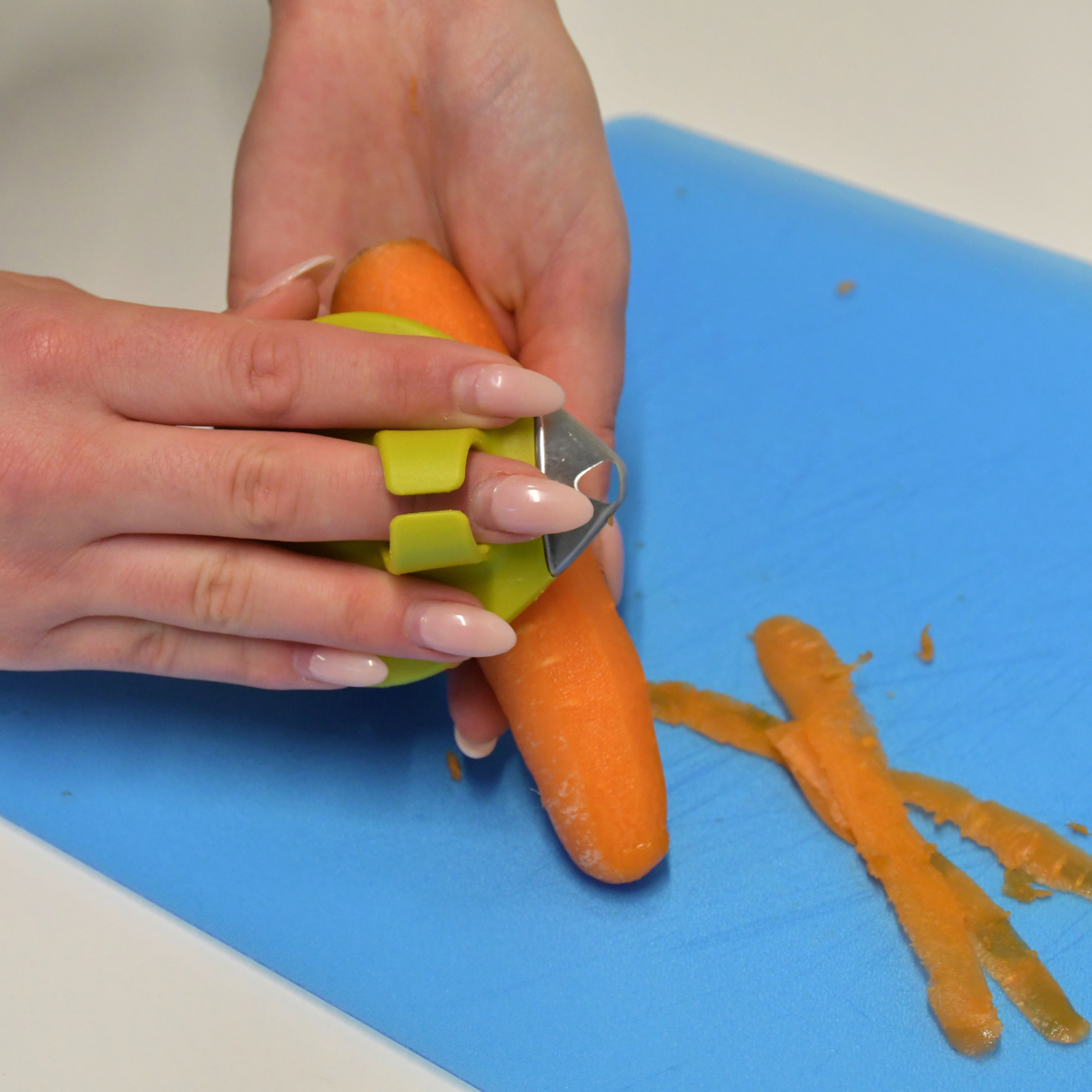 A person peeling a carrot using a bright yellow vegetable peeler over a blue chopping board.