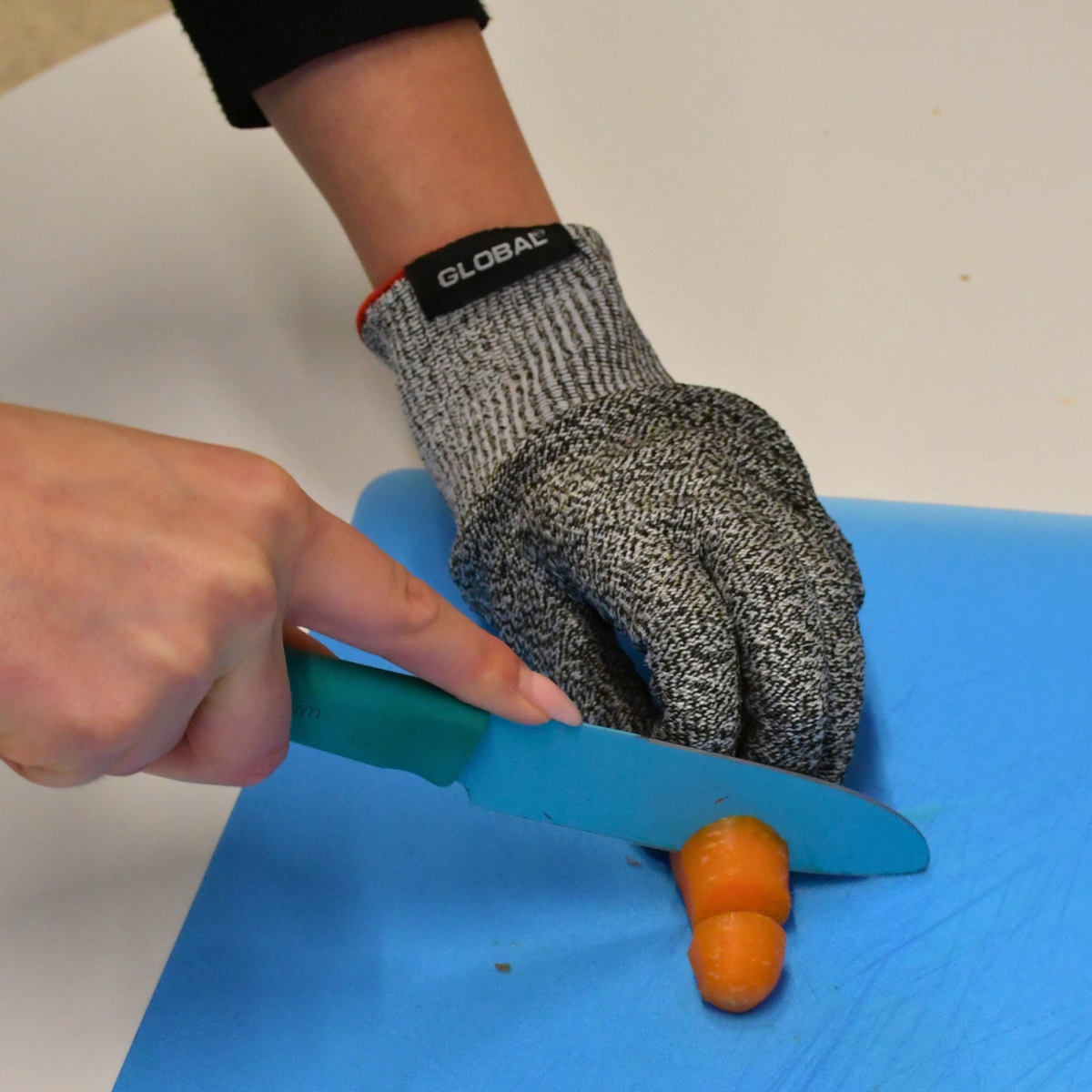 A person wearing a protective cut-resistant glove cutting a carrot on a blue chopping board.
