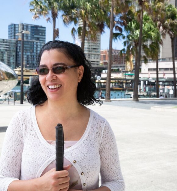 A person standing outside wearing sunglasses and holding a white cane.
