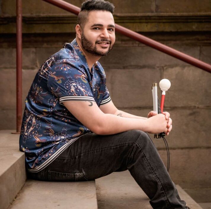Guide Dogs staff member Karan Nagrani sitting on steps outside holding his white cane.