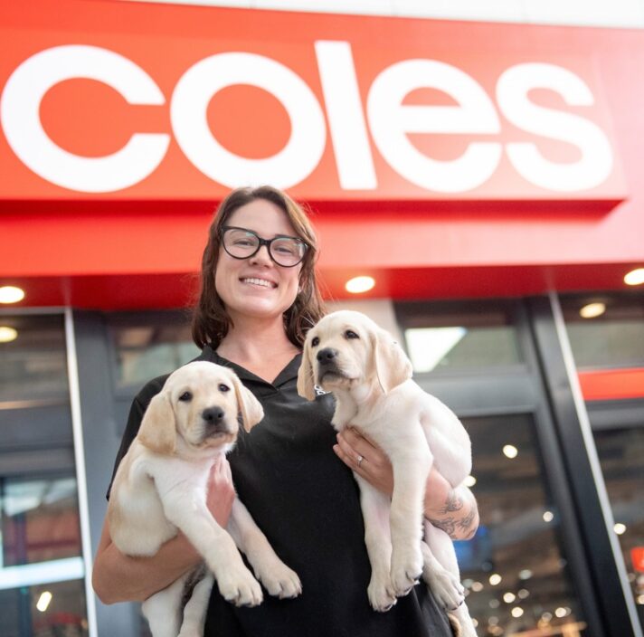 Guide Dog Trainer Chelsea holding two yellow Labradors pups outside a Coles store.