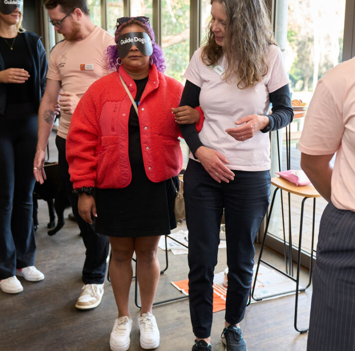 A guest wearing a red coat is being guided by an event volunteer to experience how to walk after completely losing vision.