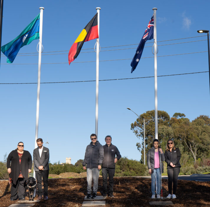 Guide Dogs Clients and staff standing in front of three flag poles with the Torres Strait Islander, Aboriginal and Australian Flags raised.