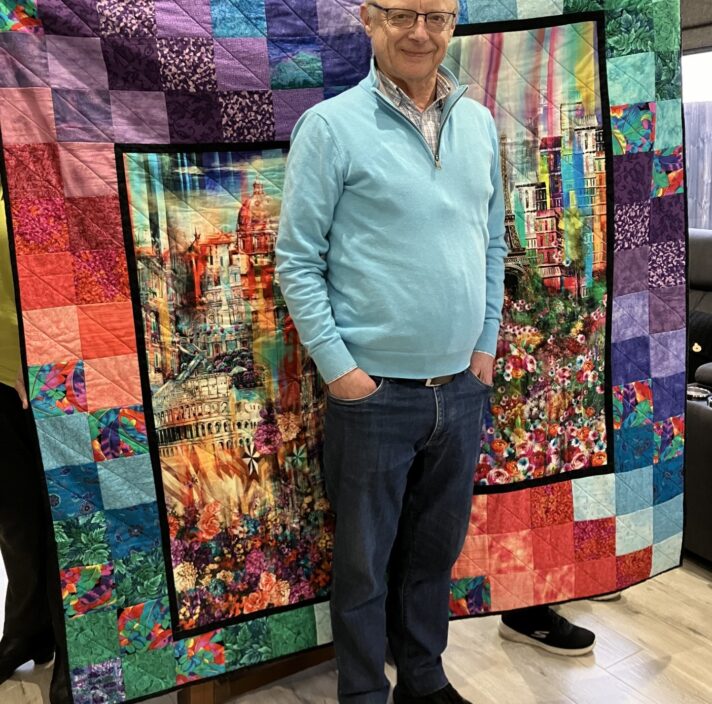 A man standing in front of a patchwork quilt, smiling at the camera.