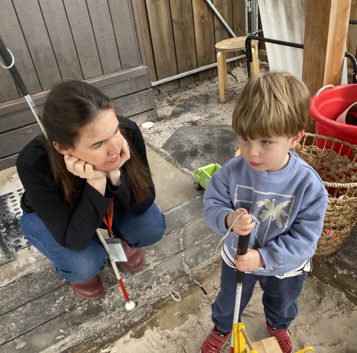 Guide Dogs staff member, Erin, kneeling down with her head in her hands, listening intently as Louie talks to her about his white cane, Sneezy.