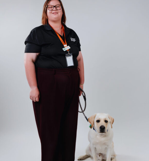 Gemma standing in her black Guide Dog uniform, smiling, as Keith, yellow ambassador Labrador, sits at her feet.