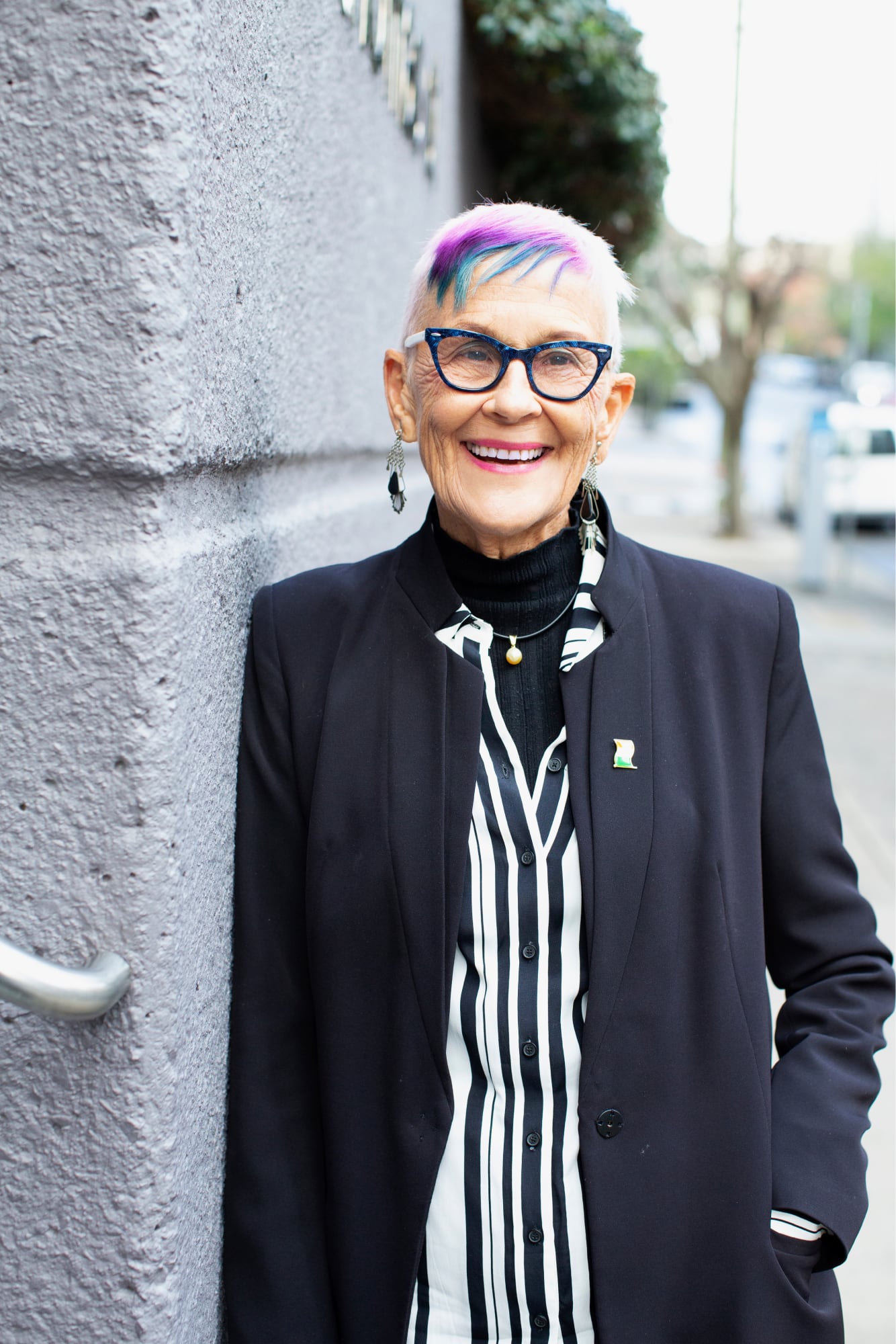 A person standing outside and leaning against a wall looking at the camera with a smile on their face.