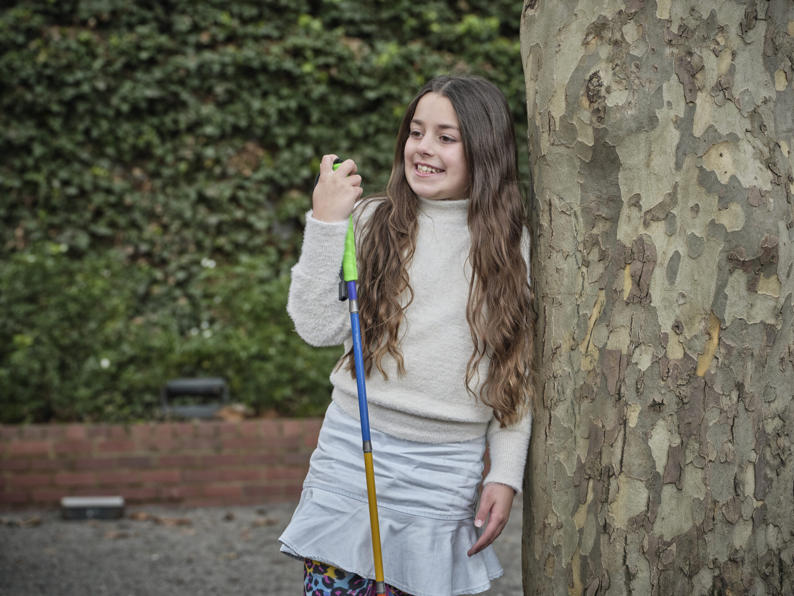 A young Client leaning against a tree, looking away smiling and holding onto her white cane.