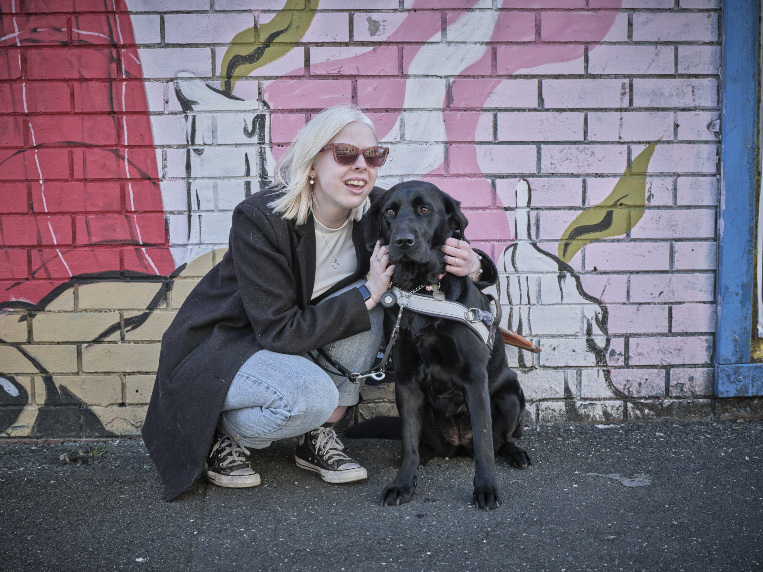 A young person kneeling down, cuddling their black Labrador Guide Dog.