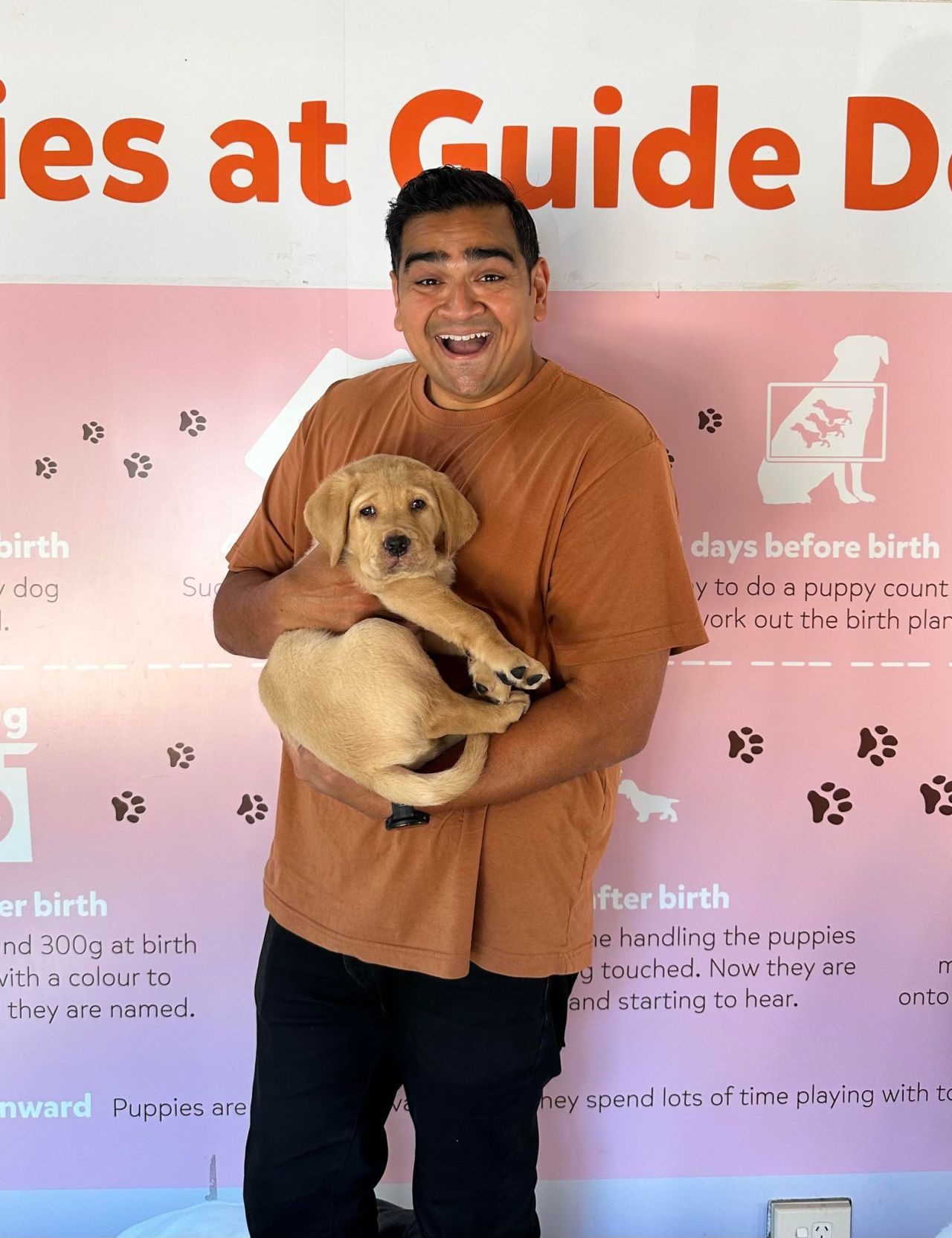 Dilruk is standing, holding a yellow Labrador puppy in his arms. He is looking at the camera with a big smile on his face.