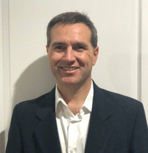 David Simmonds wears a black suit jacket with a white button down shirt. David stands in front of a beige wall, smiling and looking at the camera.