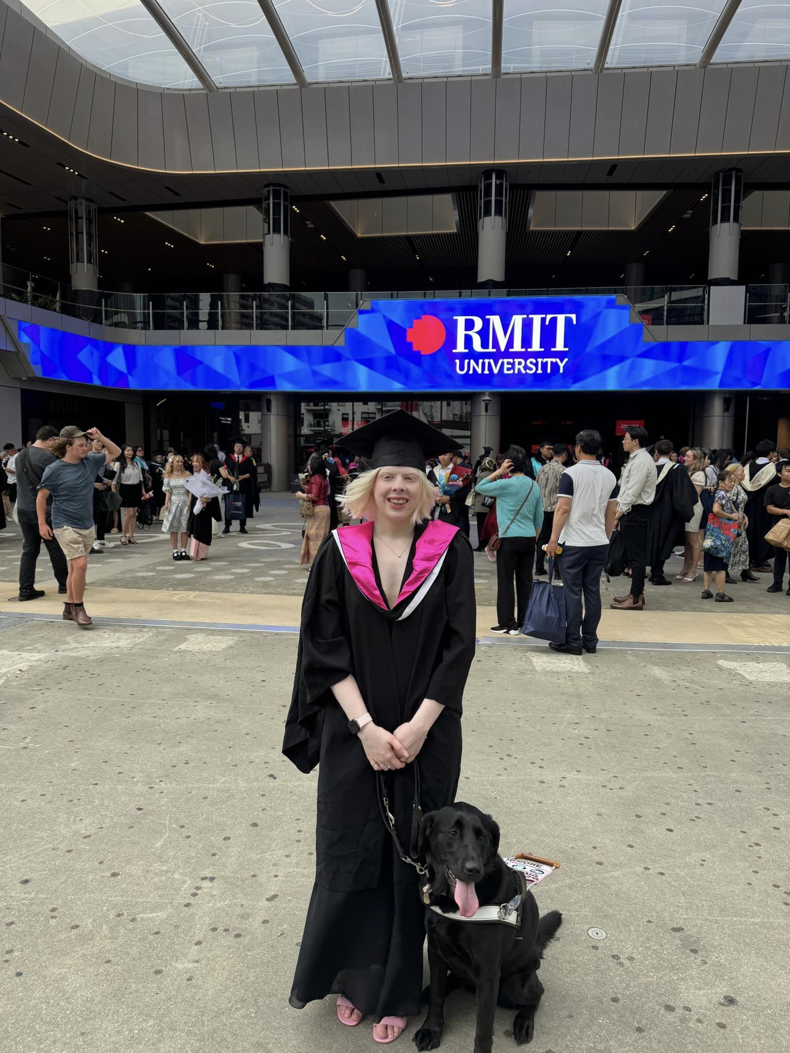 Lucy in her academic regalia featuring bright pink lining with black Labrador Dottie in harness by her side. They stand in front of a blue banner with RMIT University written in white text with people milling around in the background.