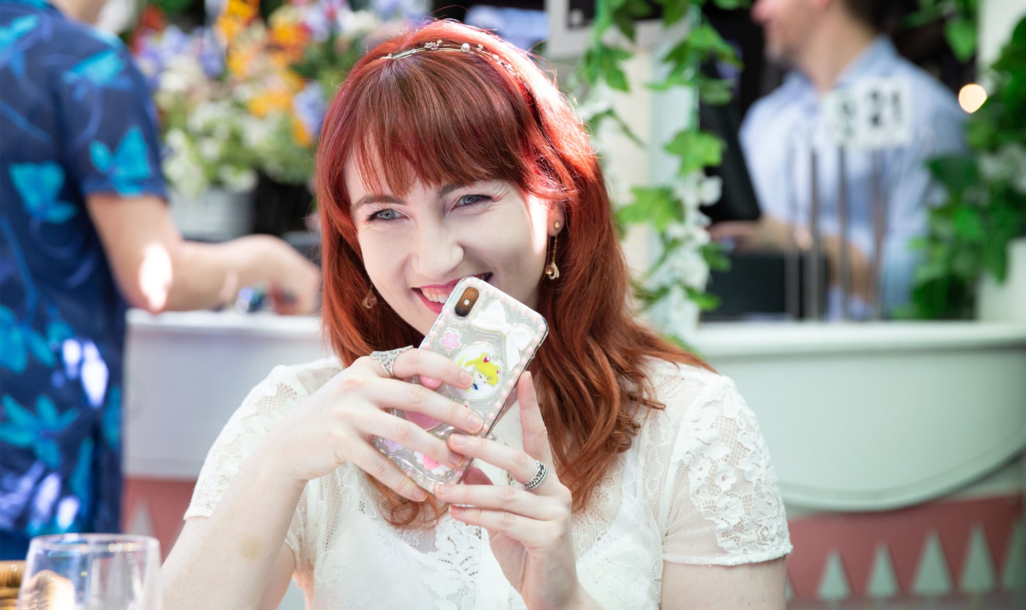 An adult who is outside at a cafe laughing. They have their phone up to their mouth and it is covering part of their mouth.