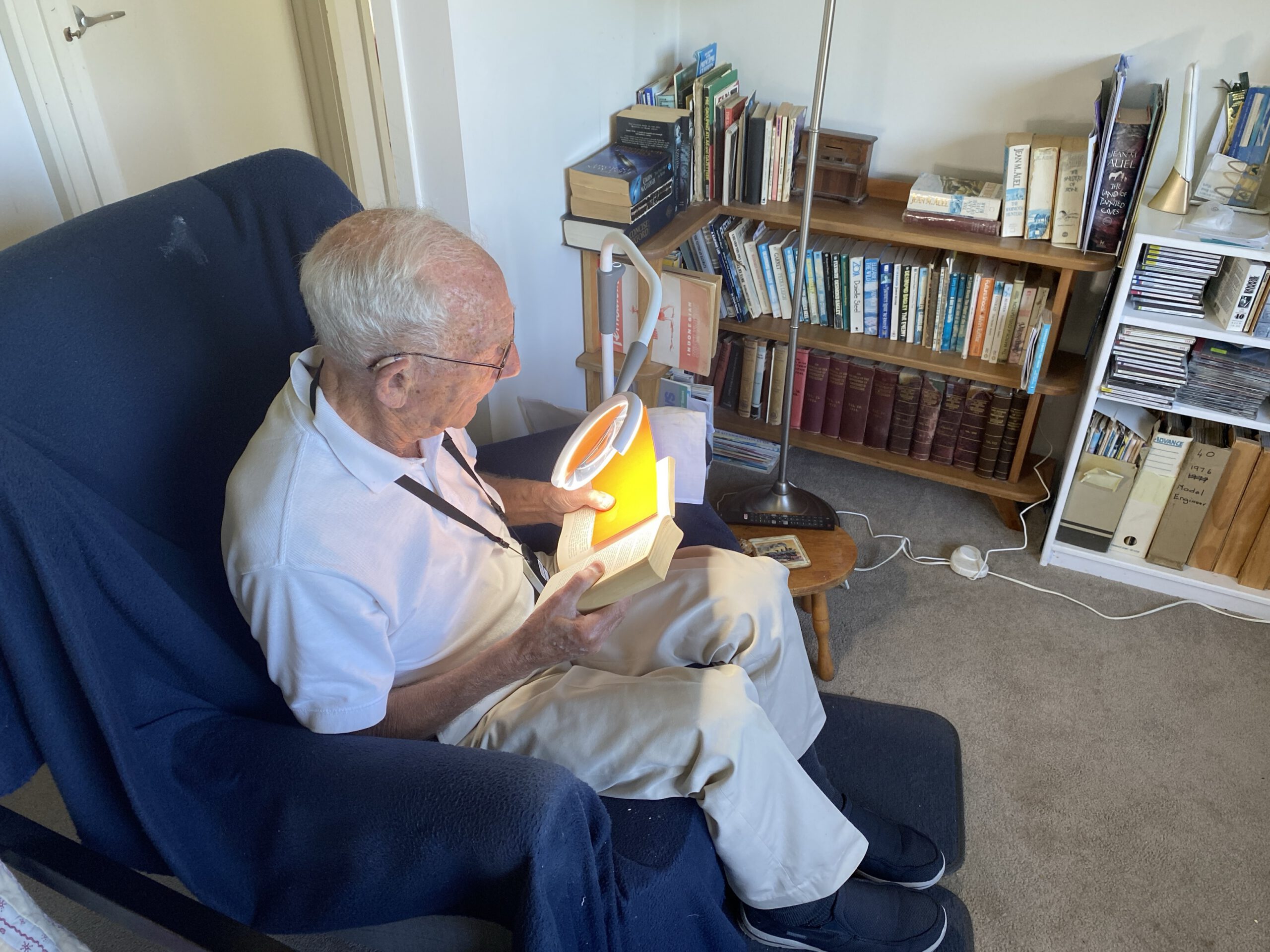 Dennis sitting in an armchair in his living room. He is reading a book using the magnifying lamp and a high contrast guide.