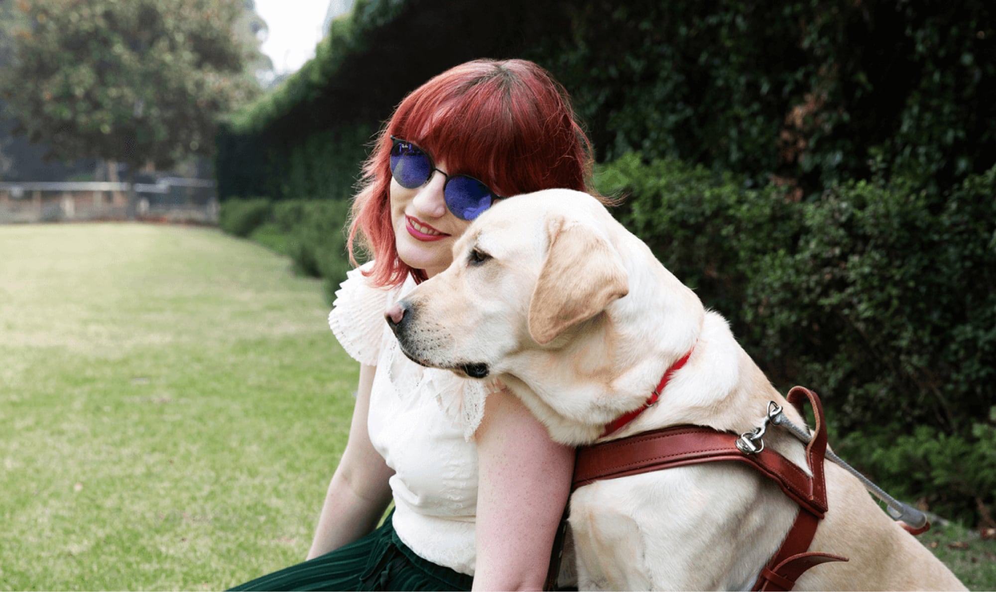 A person and a yellow Guide Dog, in harness, sitting on grass outside. The person is smiling at the camera and the Guide Dog is a profile view.