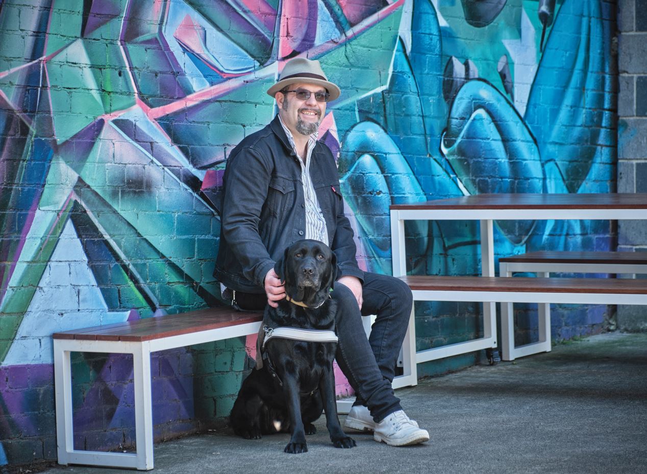 A person sitting in front of a blue graffiti wall, smiling, with their black Labrador Guide Dog sitting at their feet.