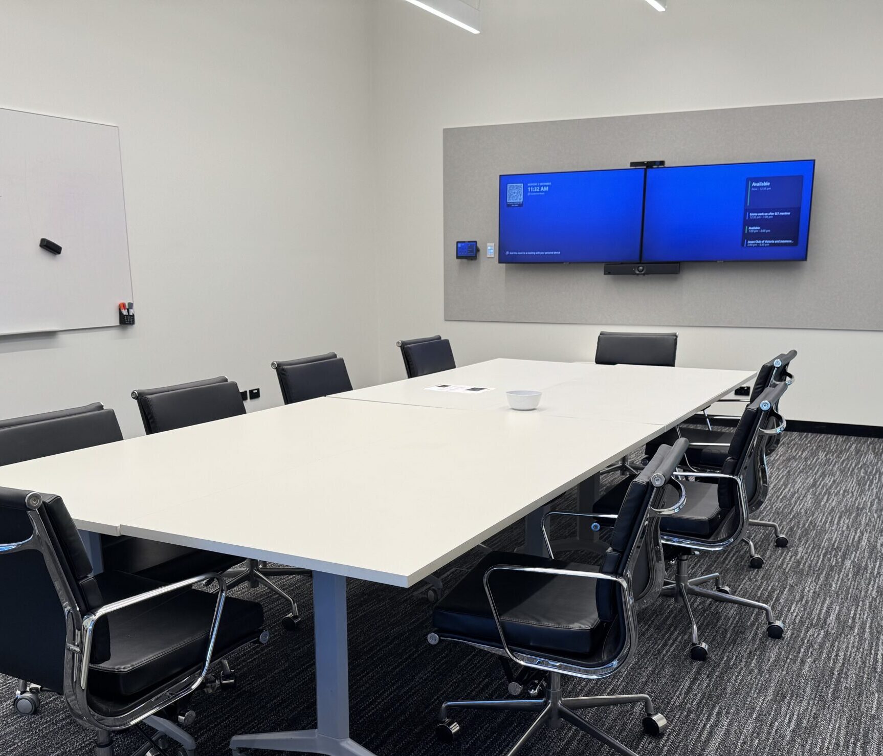 The Guide Dogs Victoria conference room with boardroom-style tables and 10 chairs set up. Two split screens are mounted on the wall displaying a blue holding screen.