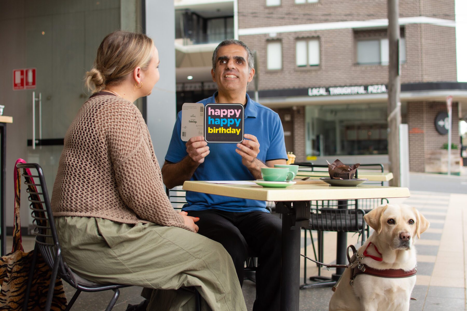 Two people at an outdoor café table, one holding a "happy happy birthday" card, with a Guide Gog beside them.