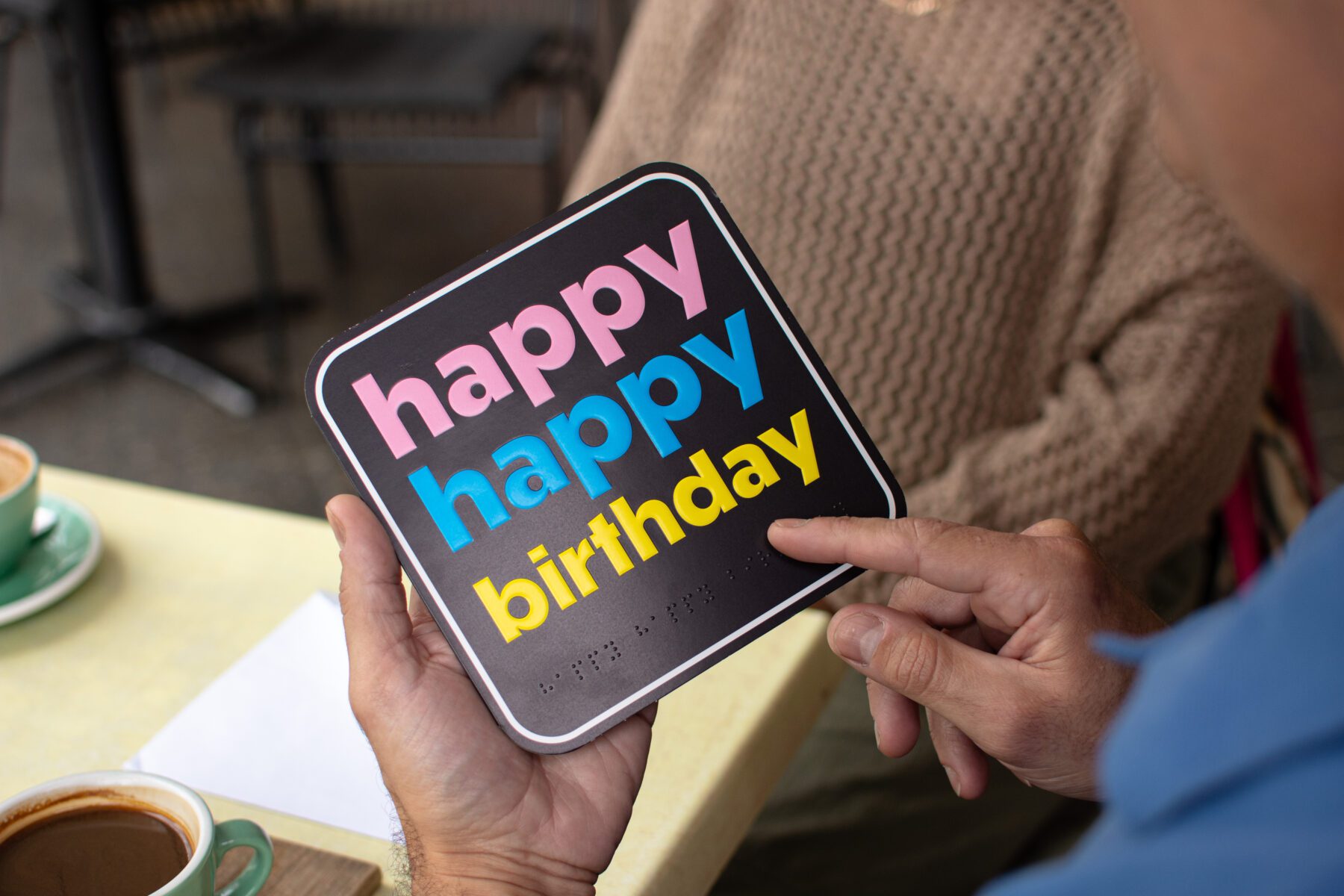 A person holding a birthday card that says "happy happy birthday" in colourful letters with braille embossed underneath the text