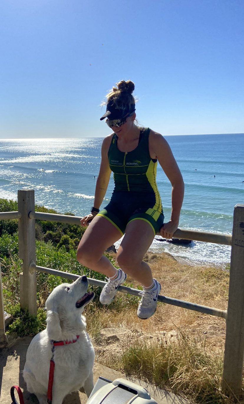 Al Viney sits on a fence at the beach in her training uniform. Charlie sits at her feet.