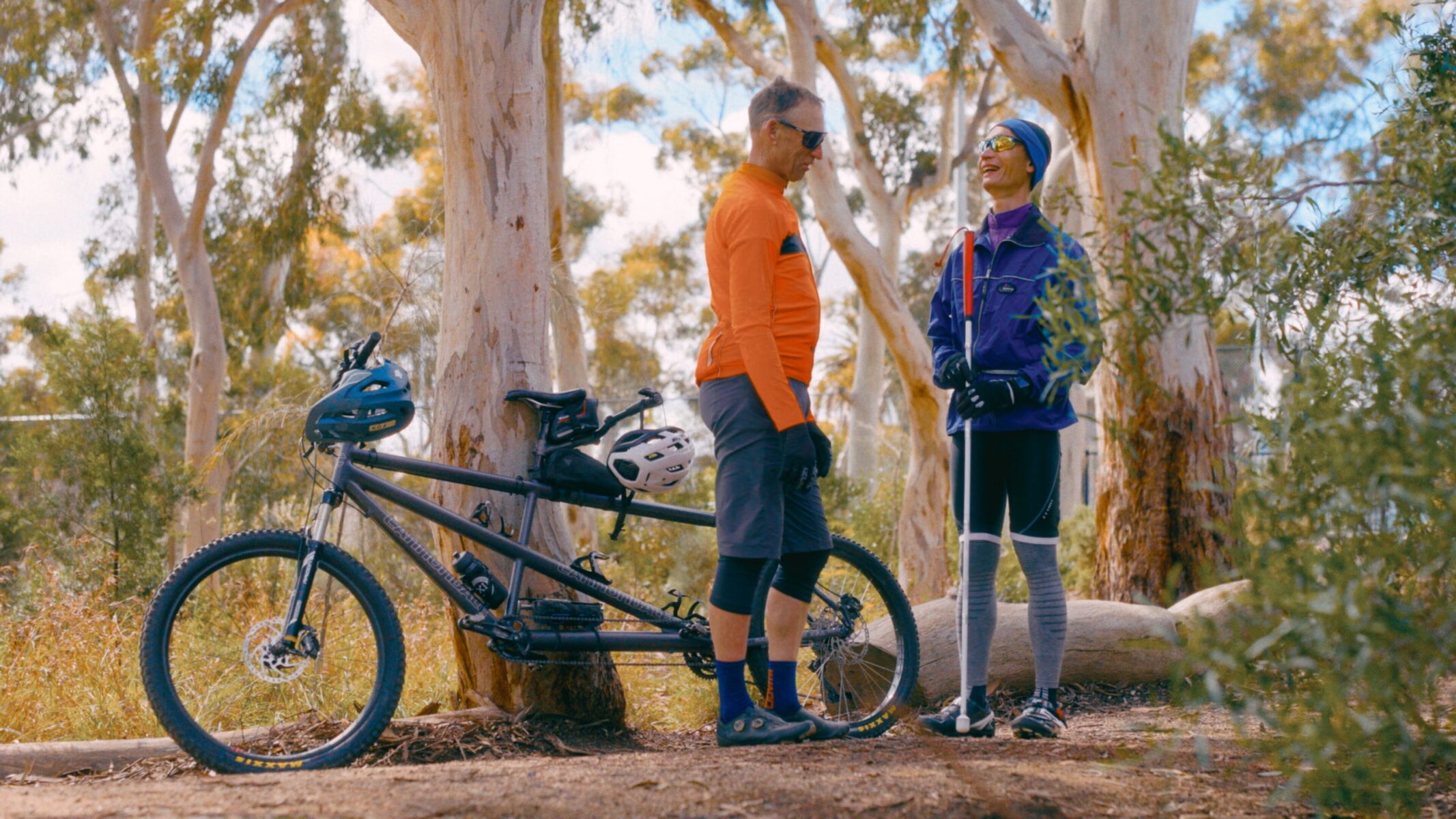Two people stand in a wooded area with tall trees and green foliage. One wears an orange long-sleeve shirt, black shorts, and blue socks, standing beside a tandem bicycle. The other wears a purple jacket and grey pants, holding a white cane. 