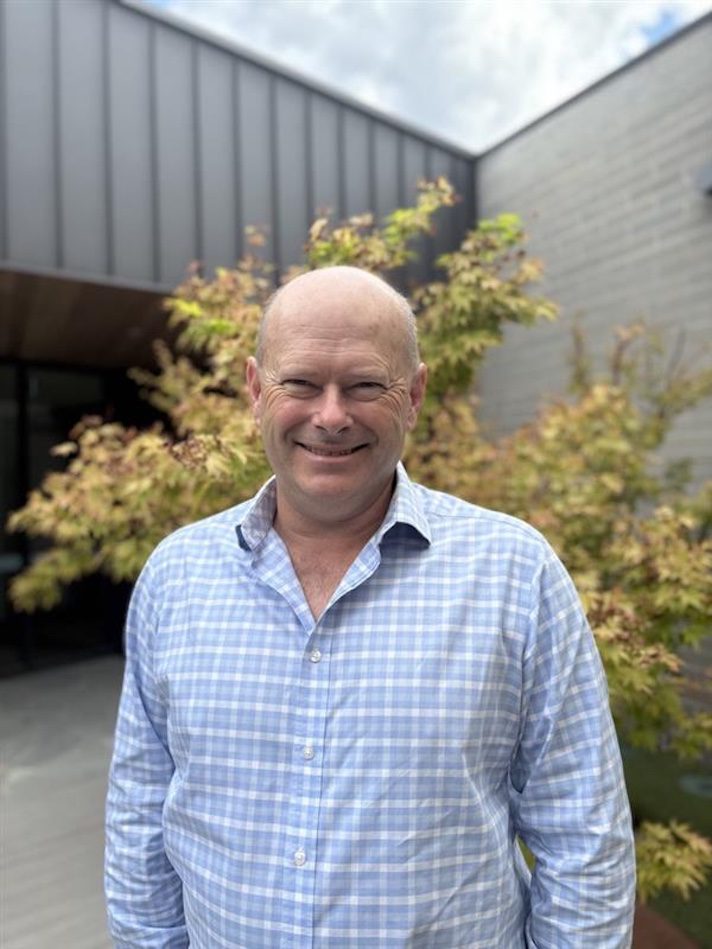 Chris McFadden wearing a light blue checkered button-up shirt stands facing the camera in an outdoor courtyard with leafy trees and modern grey buildings in the background, lit by natural daylight.