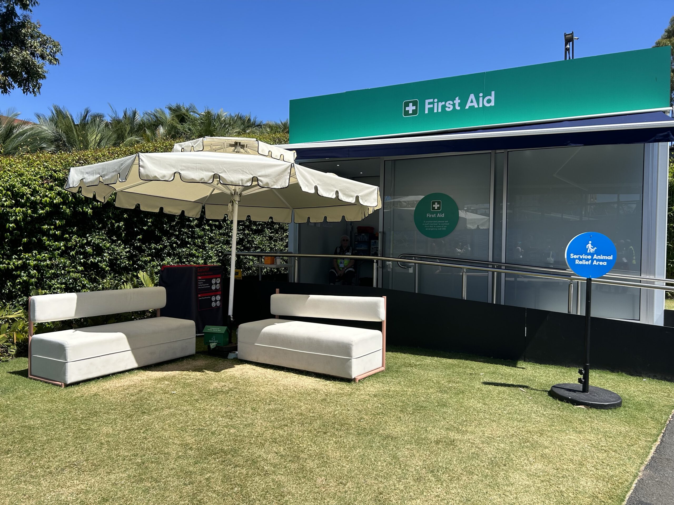 A lovely grassed area with white seats and a white umbrella, outside the First Aid building with a blue sign that says ‘Service Animal Relief Area’. 