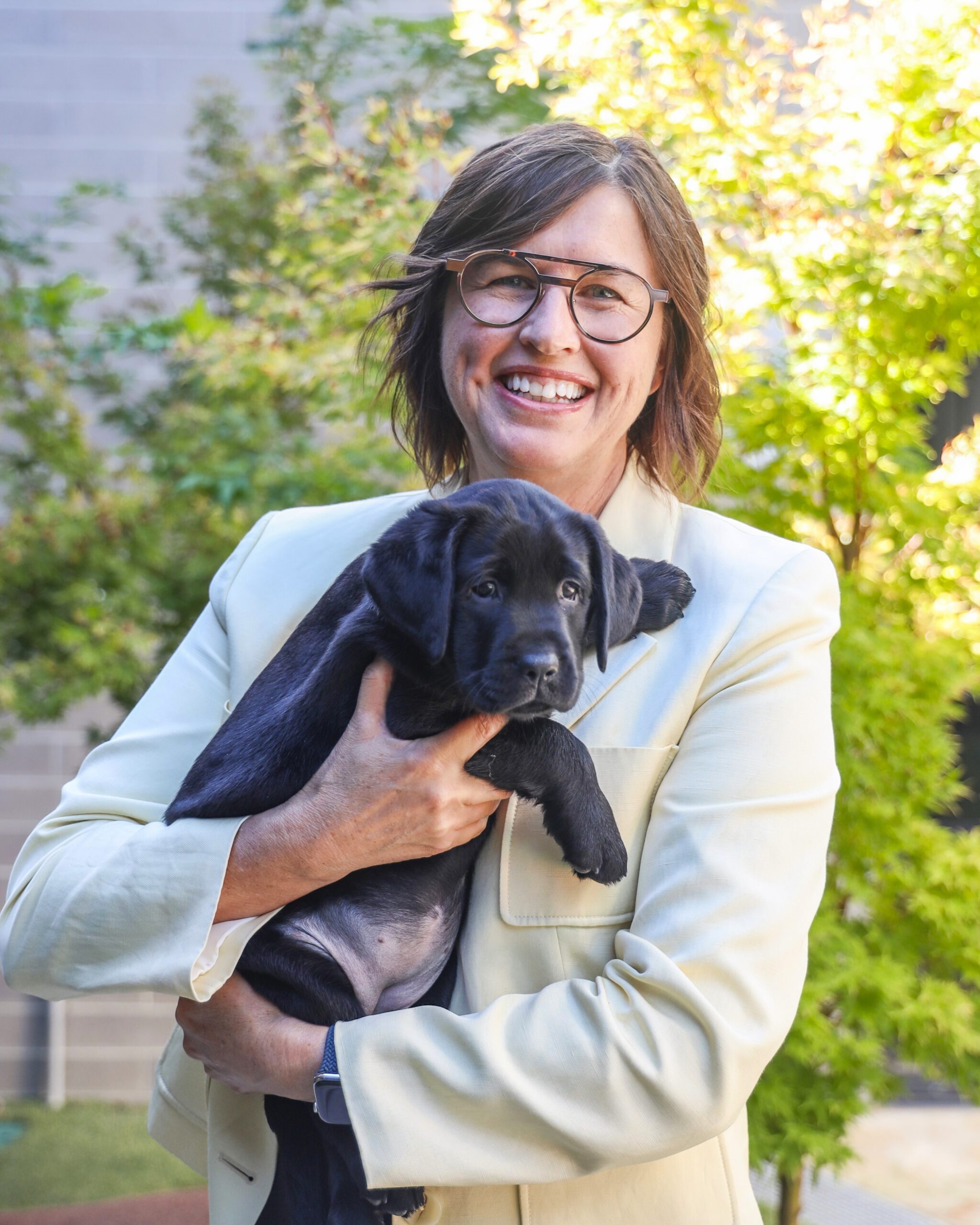 Jane Schuller holding a black Labrador puppy in her arms, smiling at the camera.