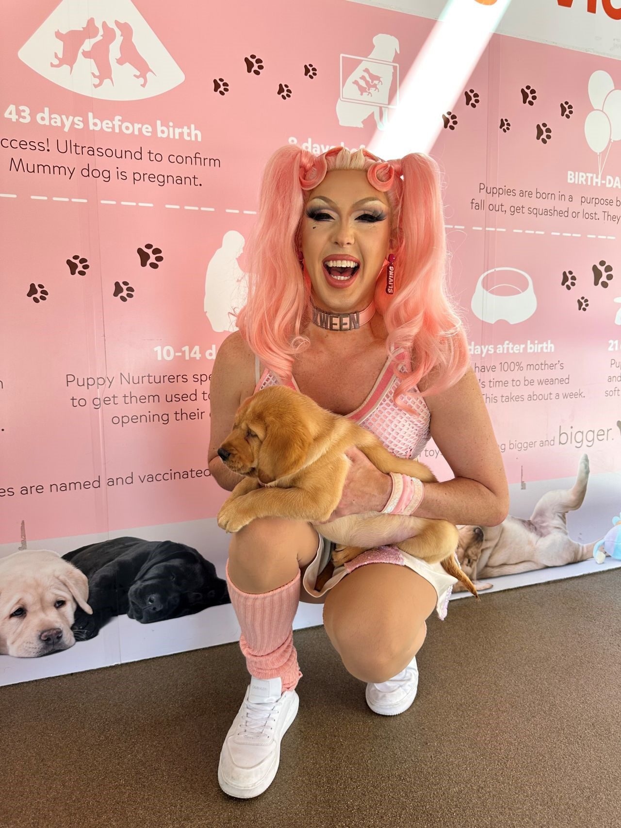 Jimi the Kween with a big smile on their face, kneeling on the ground of the Guide Dogs nursery cradling a yellow Labrador puppy.