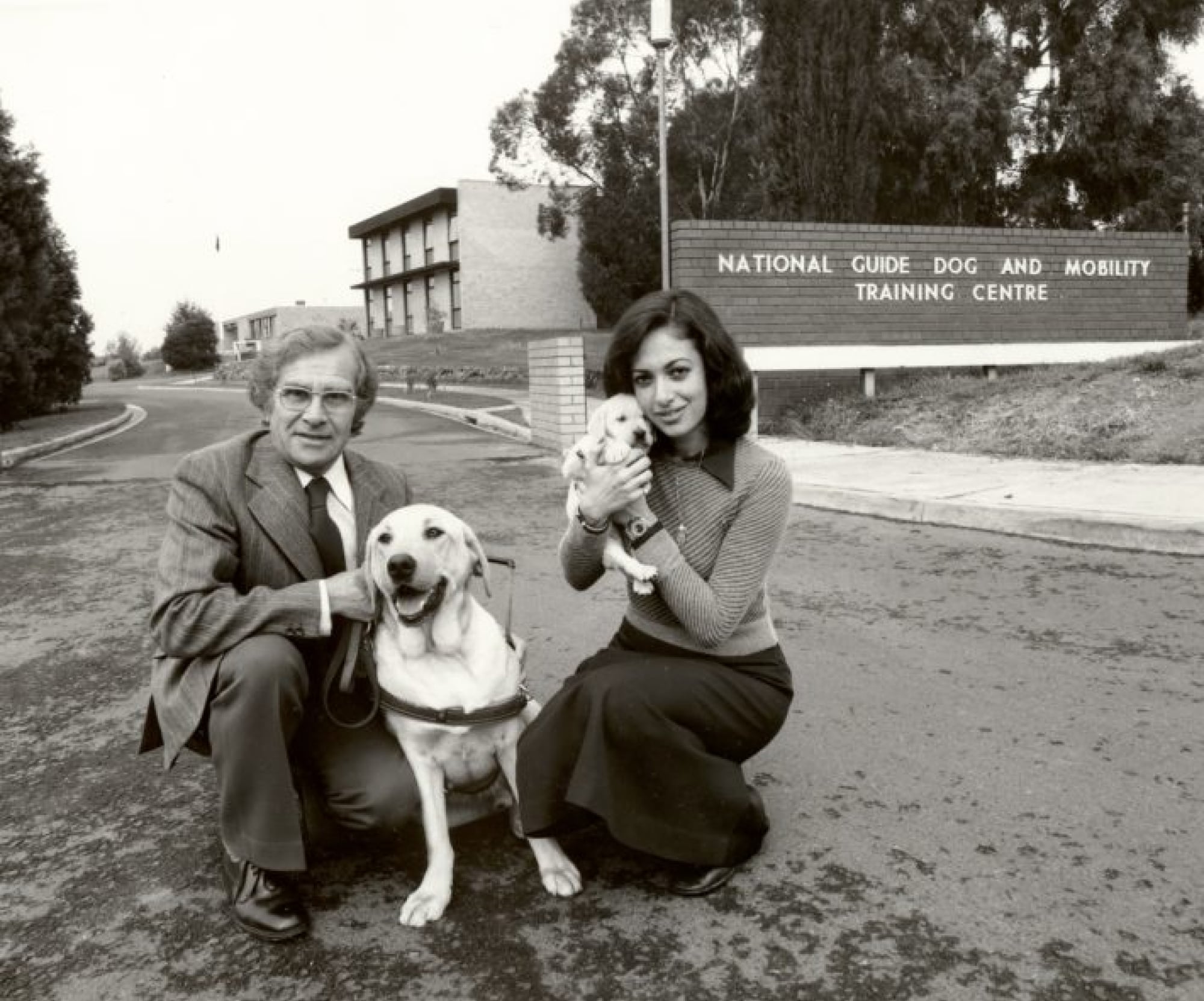 A 1940's black and white image of two people outside a Guide Dog Training Centre. One of the people in kneeling down next to a yellow labrador Guide Dog in harness and the other is holding a yellow ten week old labrador puppy. Both dogs and people are looking at the camera.
