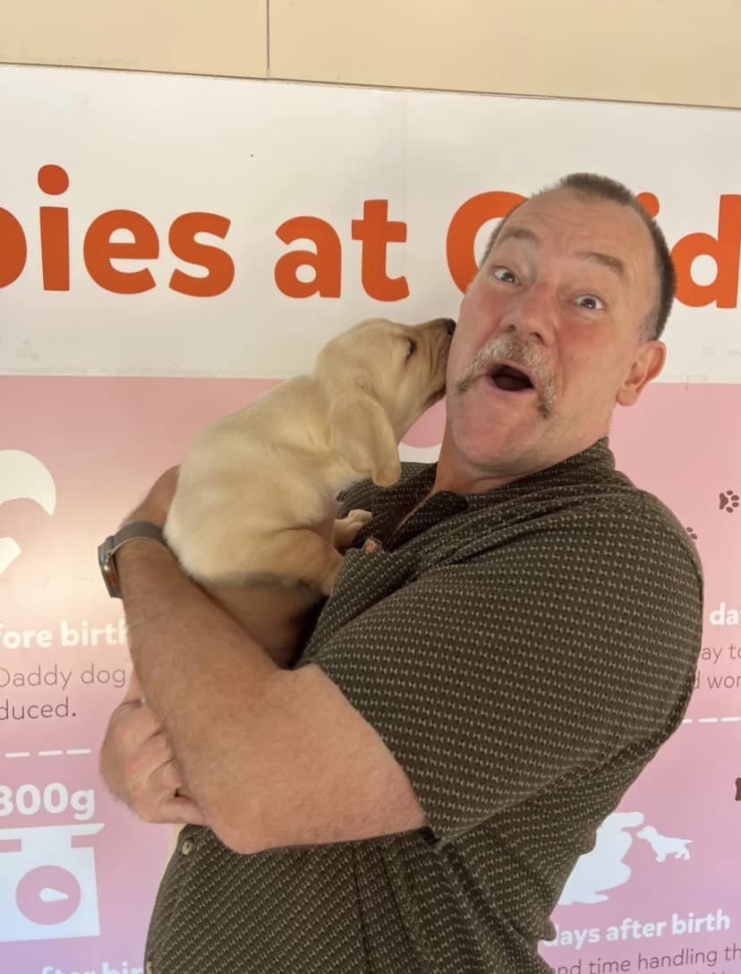 Lehmo laughing while a yellow Labrador puppy licks his ear.