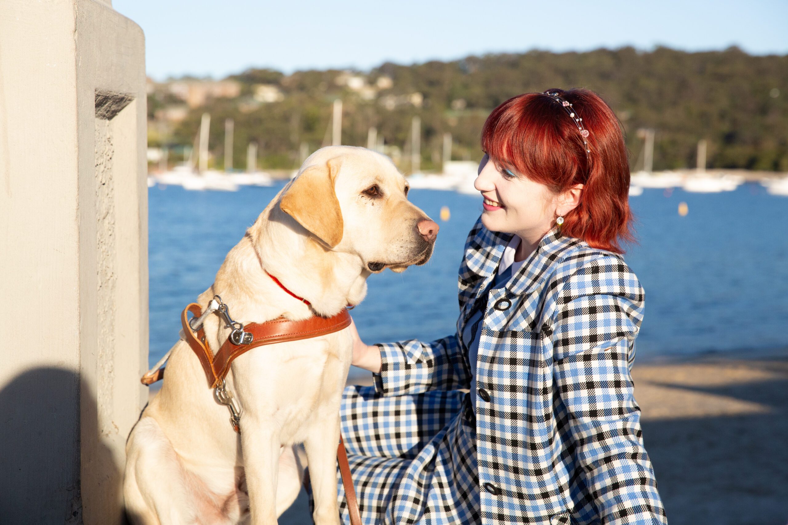 A Client smiling at her yellow Labrador Guide Dog in harness, as they sit together on a pier.