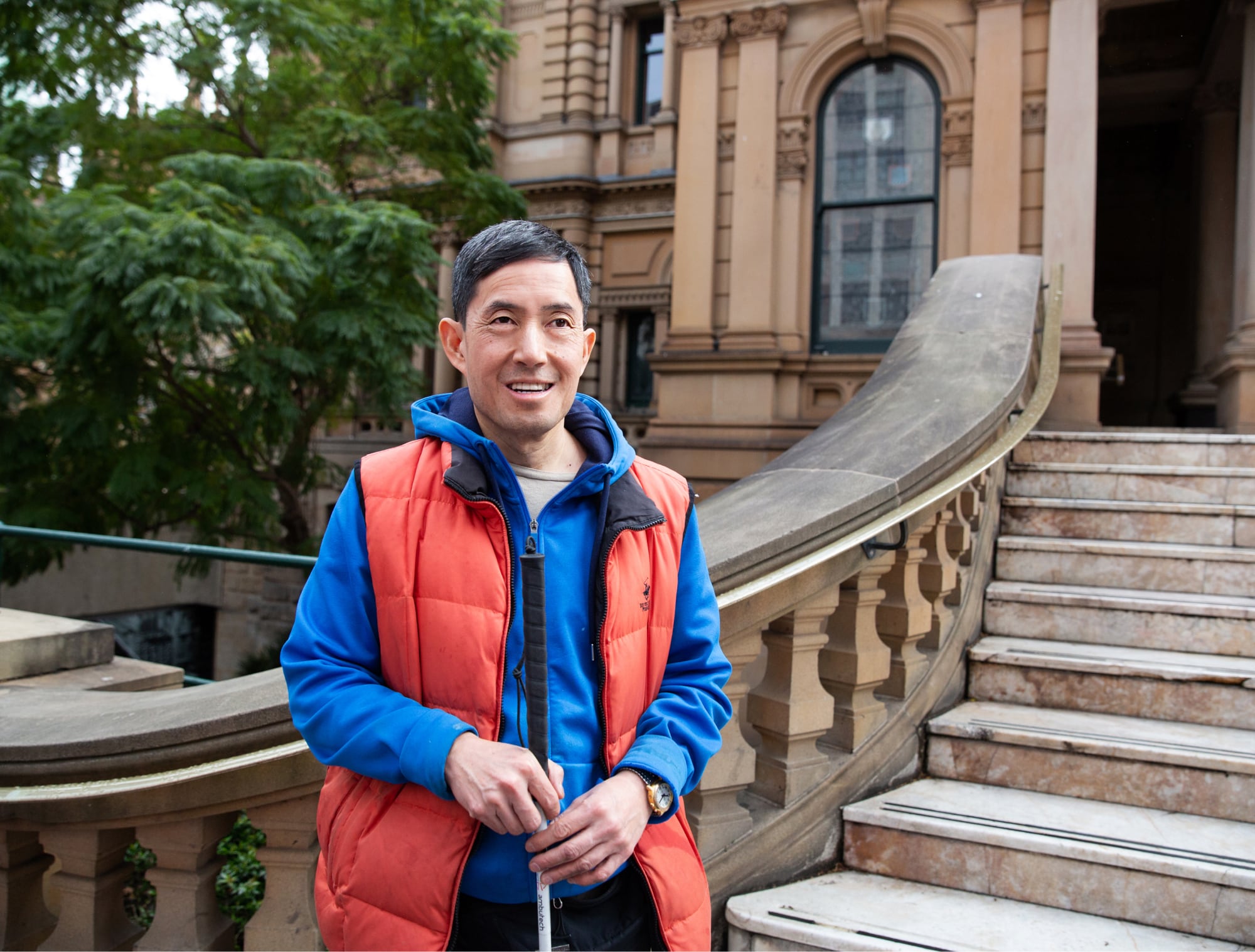 A person standing in front of a building holding their white cane. The person is smiling at the camera.