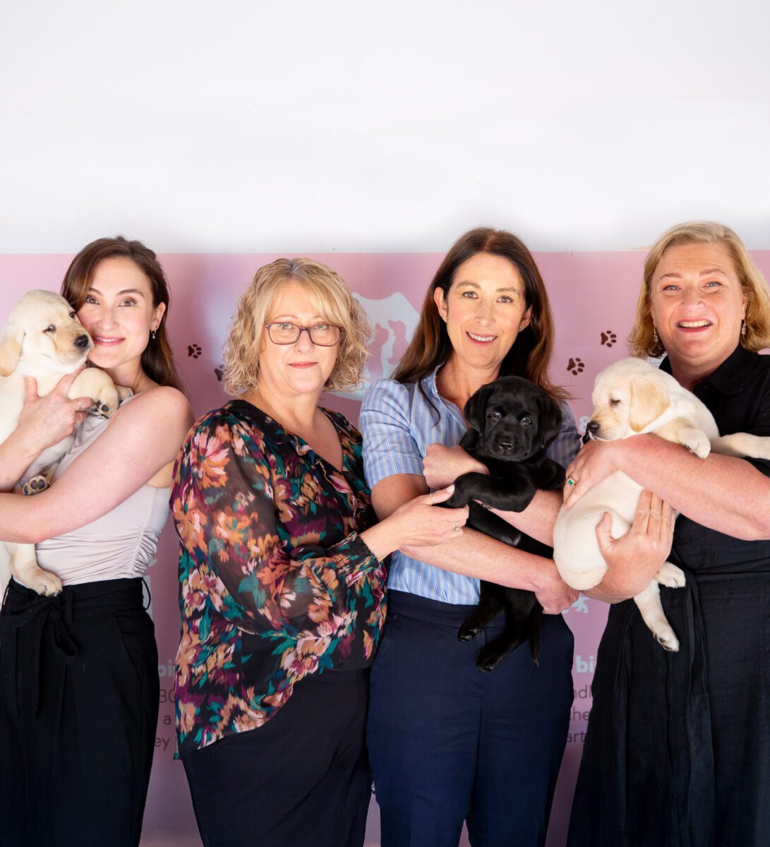 Melanie, Josie, Katie and Cath smiling at the camera holding a mix of black and yellow lab puppies.