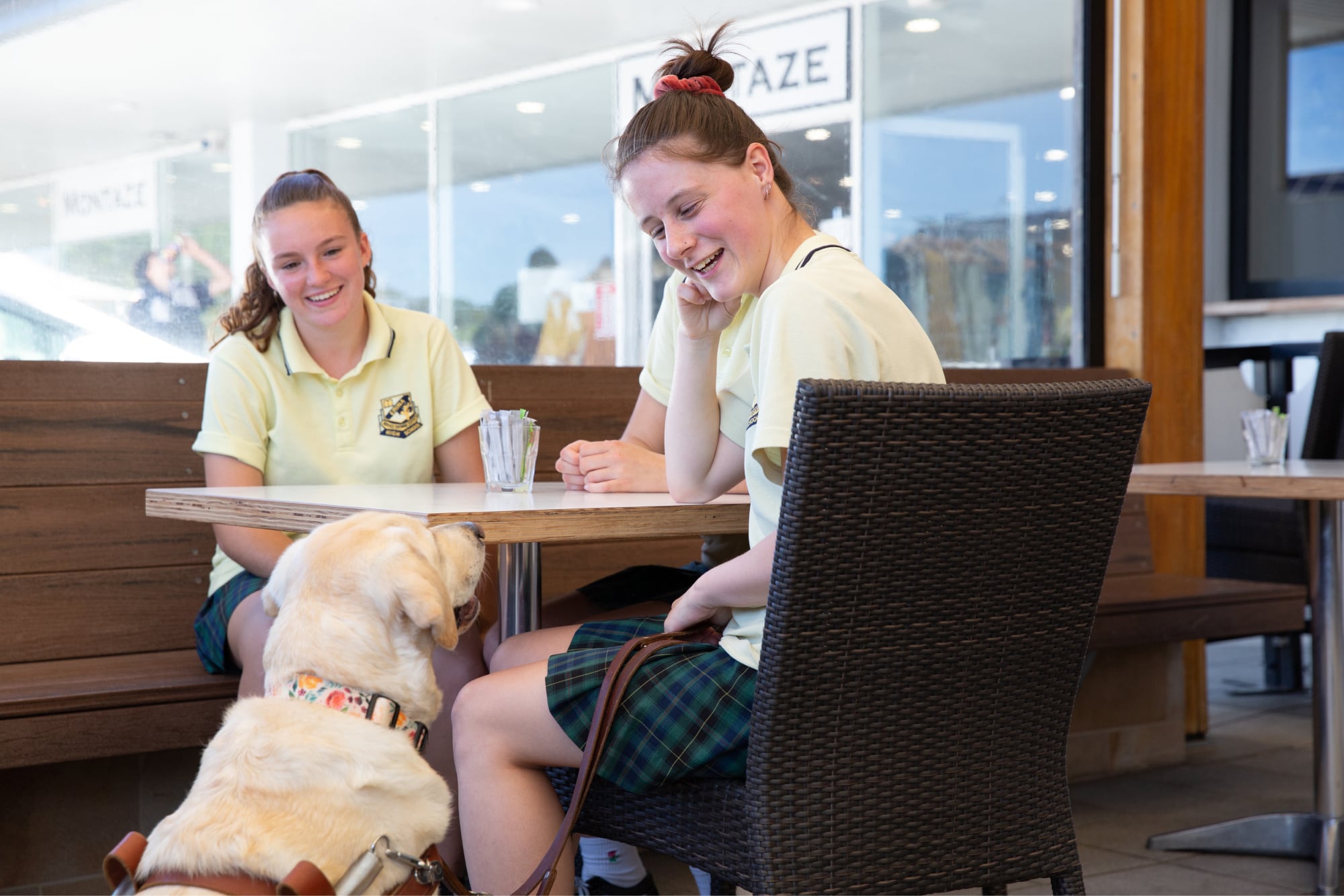 Two young adults sitting a cafe table. A yellow Guide Dog in harness is seated next to the table looking up at the people. The young adults are looking down at the Guide Dog smiling.