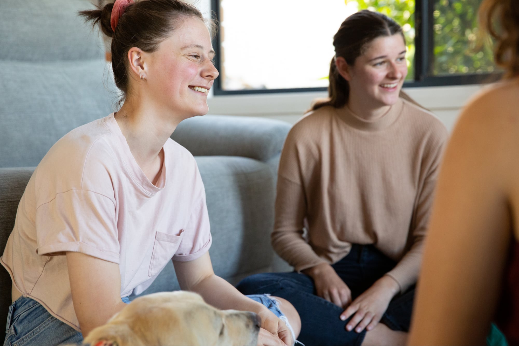 Three young adults sitting inside with a yellow labrador dog next to one of them. They are all looking at each other smiling.