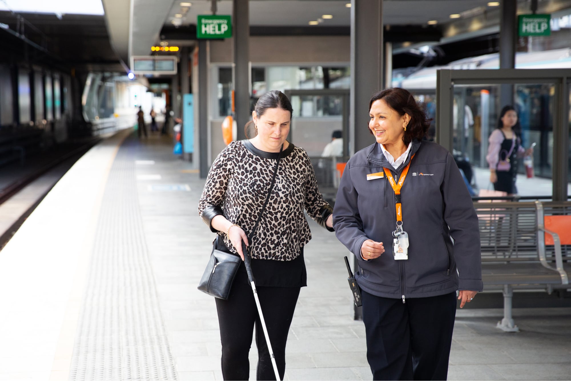 A person using their white cane at the train station while being guided by a train station attendant.