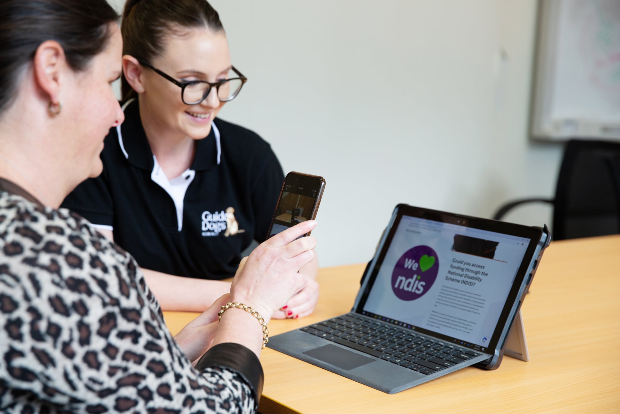 A person and a Guide Dog's staff member looking at a laptop. The person is holding up an Iphone to the laptop and the staff member is smiling.